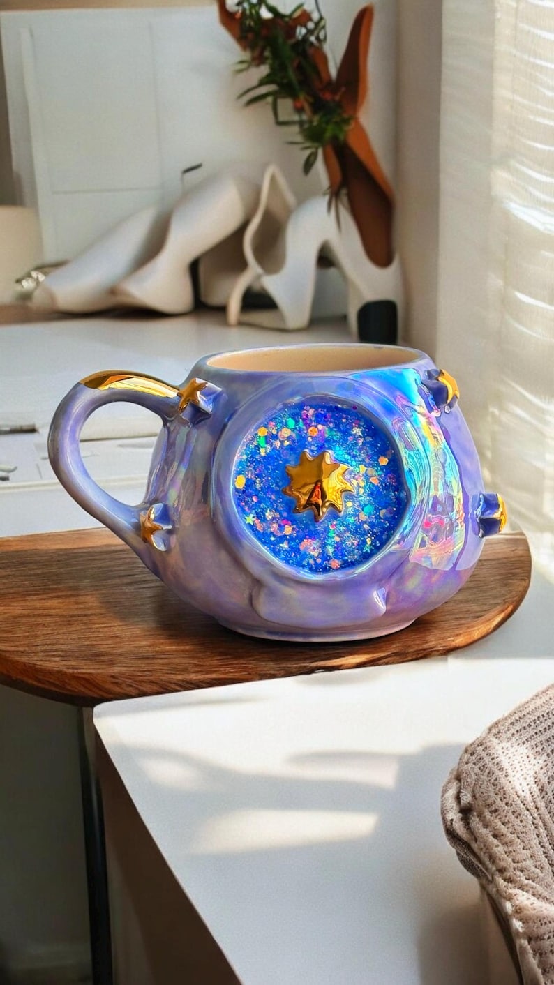 May include: A ceramic mug with a celestial theme. The mug is a lavender color with gold accents and a large, round window filled with blue glitter and a gold star. The handle and star details are gold. The mug sits on a wooden surface.