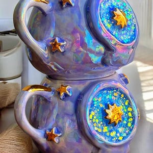 May include: Iridescent ceramic mug with a celestial theme. The mug is lavender with gold star accents and a gold handle. The design includes a blue and gold starburst pattern. The mug is a unique piece of drinkware.