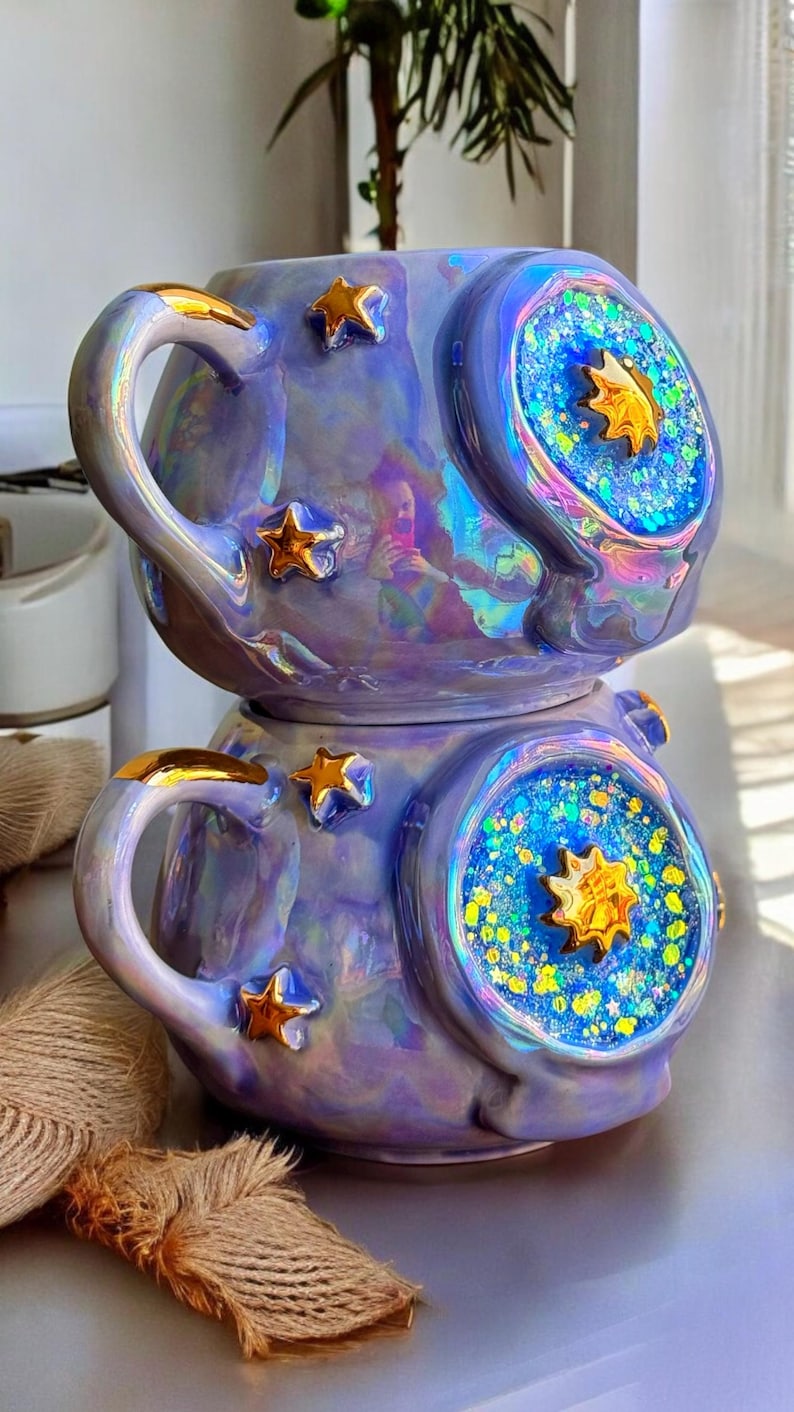 May include: Iridescent ceramic mug with a celestial theme. The mug is lavender with gold star accents and a gold handle. The design includes a blue and gold starburst pattern. The mug is a unique piece of drinkware.