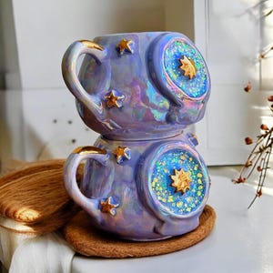 May include: Two stacked, iridescent purple ceramic mugs with gold star accents. Each mug features a circular design with a gold star in the center, surrounded by blue and green glitter. The handles are trimmed with gold.