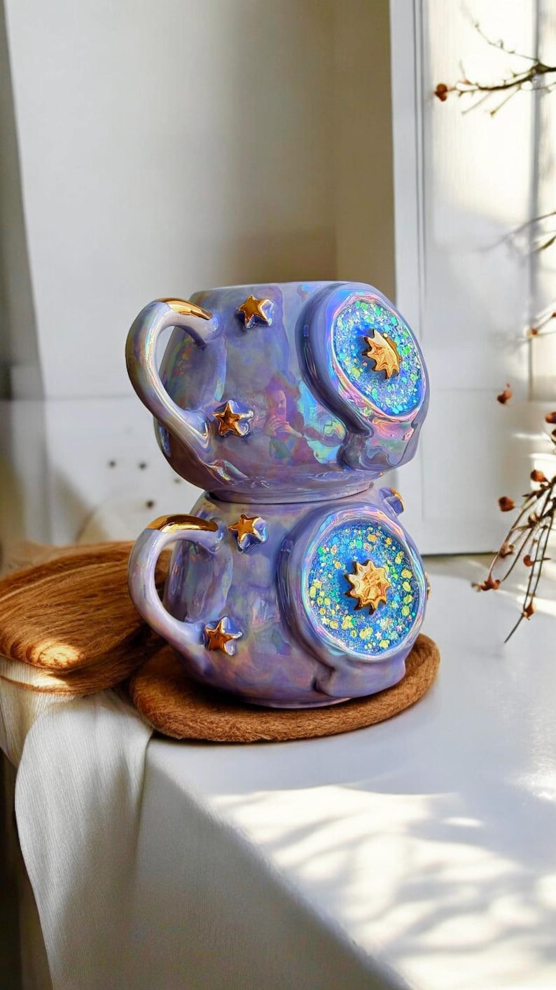 May include: Two stacked, iridescent purple ceramic mugs with gold star accents. Each mug features a circular design with a gold star in the center, surrounded by blue and green glitter. The handles are trimmed with gold.