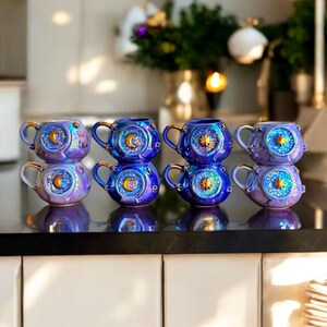 May include: A collection of eight iridescent ceramic mugs with celestial designs. Each mug features a blue and purple color scheme, gold handles, and a sun, moon, and star motif. The mugs are stacked in pairs on a dark countertop.