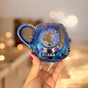 Puede incluir: Una taza de cerámica azul iridiscente con un diseño de media luna dorada y purpurina. La taza tiene un asa y el nombre "DIANA" está impreso en la parte delantera. La taza es sostenida por una persona.