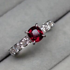 Könnte beinhalten: Ein silberner Ring mit einem großen, quadratisch geschliffenen, tiefroten Edelstein in der Mitte. Das Band ist mit kleineren, klaren Edelsteinen besetzt. Der Ring wird vor einem weichen, grauen Hintergrund präsentiert, der das Funkeln der Steine hervorhebt.