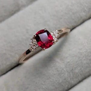 May include: A gold ring featuring a large, rectangular, deep red gemstone as the focal point. Small, clear accent stones are clustered on either side of the main stone. The ring's band is a delicate gold color.