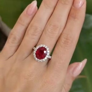 May include: A ruby and diamond ring. The ring features a large, oval-cut, deep red ruby surrounded by a halo of smaller, round, clear diamonds. The band is a polished silver color. The ring is displayed on a hand.