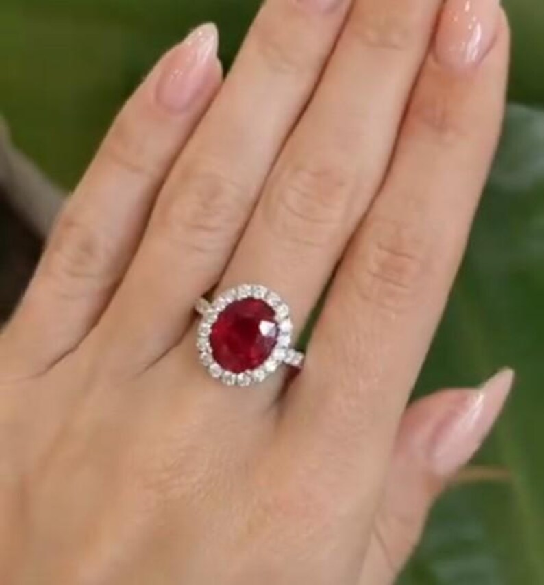 May include: A ruby and diamond ring. The ring features a large, oval-cut, deep red ruby surrounded by a halo of smaller, round, clear diamonds. The band is a polished silver color. The ring is displayed on a hand.
