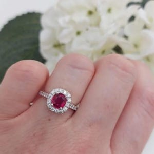 Könnte beinhalten: Ein silberner Ring mit einem großen, ovalen, rubinroten Edelstein, umgeben von einem Kranz aus kleineren, klaren Edelsteinen. Der Ring befindet sich an einer Hand, mit einem verschwommenen Hintergrund aus weißen Blumen.