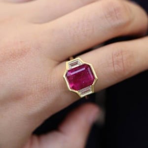 Op de afbeelding: Een gouden ring met een grote, rechthoekige, dieprode edelsteen als middelpunt. Twee kleinere, rechthoekige, heldere edelstenen flankeren de hoofdsteen. De zetting van de ring heeft een geometrisch ontwerp.
