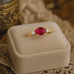 May include: A gold ring featuring a central oval-cut red gemstone flanked by two smaller round-cut clear stones. The ring is set in a polished yellow gold band with a prong setting, displayed on a beige velvet ring box.