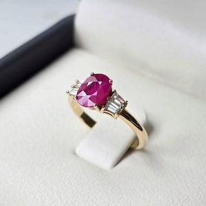 May include: A gold ring with a large, oval ruby as the central stone, and two smaller, rectangular diamonds on either side. The ring is displayed on a white stand inside a black and white box.