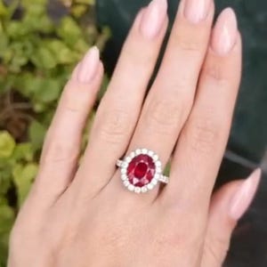 May include: A silver ring with a large, oval-cut, deep red gemstone surrounded by a halo of smaller, clear stones. The ring is set on a hand with manicured nails, with a blurred green background.