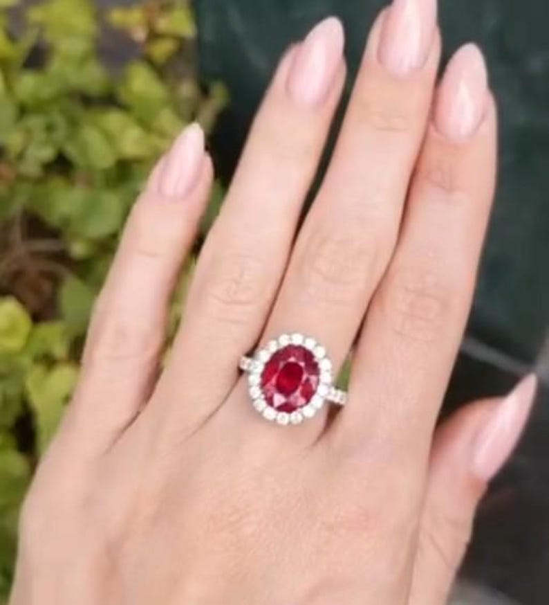 May include: A silver ring with a large, oval-cut, deep red gemstone surrounded by a halo of smaller, clear stones. The ring is set on a hand with manicured nails, with a blurred green background.