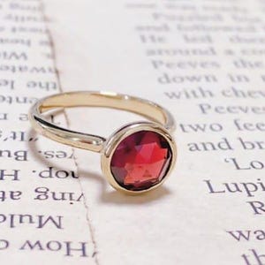 Peut inclure: Une bague dorée ornée d'une pierre précieuse rouge foncé, ronde et facettée. La bague est posée sur une page de livre vintage avec du texte visible. La pierre précieuse est le point central, captant la lumière.