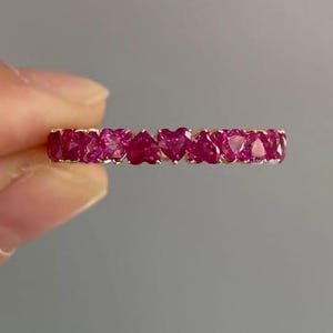 May include: A gold ring featuring a series of heart-shaped, vibrant red gemstones. The gemstones are set closely together, creating a continuous band of color. The ring is held against a neutral gray background.