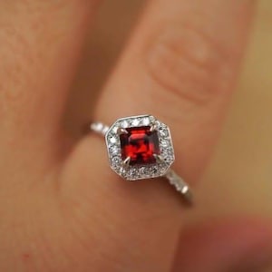 Op de afbeelding: Een zilveren ring met een vierkant geslepen, dieprode edelsteen, omringd door een halo van kleinere, heldere stenen. De ringband heeft kleine, sprankelende accenten. De ring wordt getoond op een vinger, met een vage achtergrond.