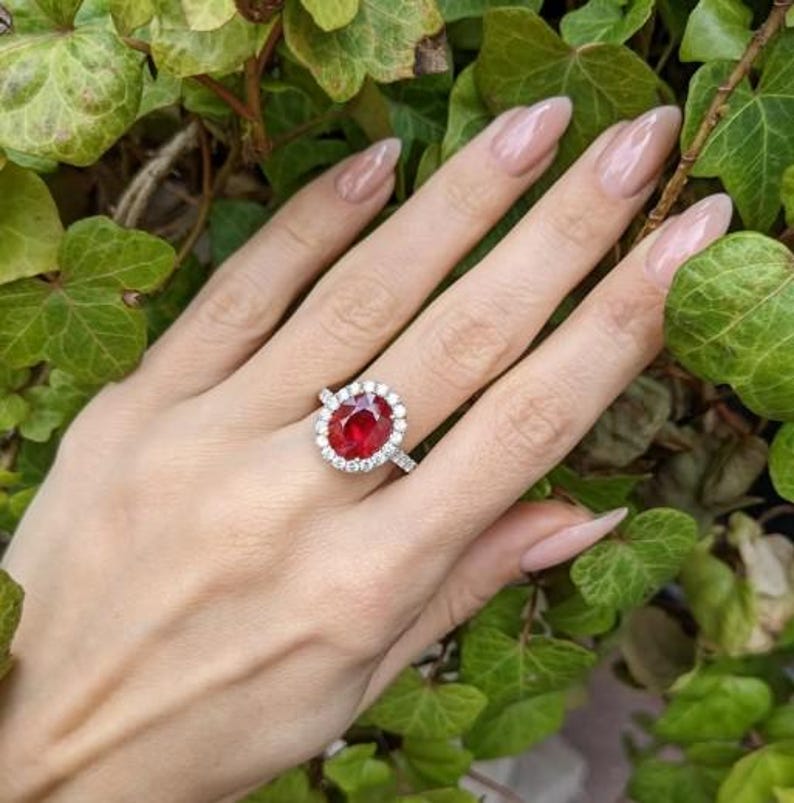 May include: A silver ring with a large oval-cut ruby center stone, surrounded by a halo of small, round diamonds. The ring is set on a delicate band. The hand is surrounded by green leaves.