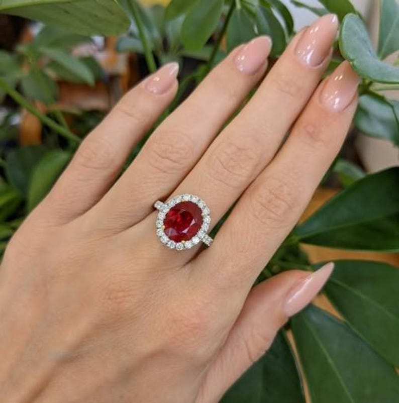 May include: A hand wearing a silver ring with a large, oval-cut, deep red gemstone. The gemstone is surrounded by a halo of smaller, clear stones. The ring is set against a background of green leaves.