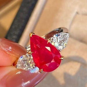 Pode incluir: Um anel com uma grande pedra vermelha vibrante em forma de pêra como ponto focal, ladeada por duas pedras menores, claras, semelhantes a diamantes. O anel tem uma cravação em ouro e uma banda prateada, mostrando um design luxuoso.