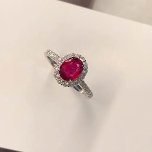 May include: A silver ring featuring a large, oval-cut ruby gemstone surrounded by a halo of smaller, clear gemstones. The ring's band is also adorned with small, clear gemstones. The ruby is a deep, vibrant red color.