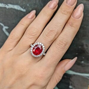 May include: A silver ring featuring a large, oval-cut, vibrant red gemstone surrounded by a halo of smaller, clear stones. The ring is set on a hand with manicured nails, showcasing the jewelry's brilliance and design.