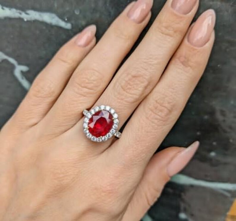 May include: A silver ring featuring a large, oval-cut, vibrant red gemstone surrounded by a halo of smaller, clear stones. The ring is set on a hand with manicured nails, showcasing the jewelry's brilliance and design.