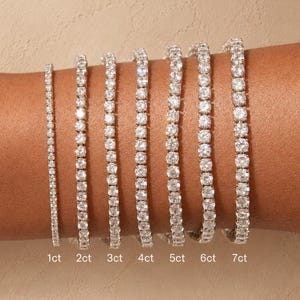 Puede incluir: Siete pulseras de tenis plateadas con piedras redondas transparentes de varios tamaños, etiquetadas como 1ct, 2ct, 3ct, 4ct, 5ct, 6ct y 7ct. Cada pulsera muestra una fila continua de piedras engastadas en una banda metálica, ordenadas por tamaño.