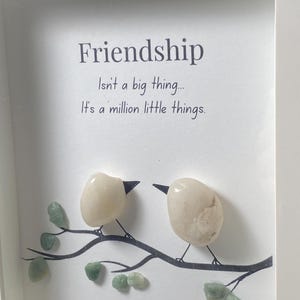 May include: Framed artwork with the word "Friendship" and a quote. Two birds made from smooth, light-coloured stones sit on a branch with green stone leaves. The artwork is set against a white background.