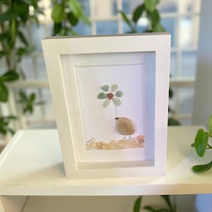 Puede incluir: Una obra de arte enmarcada con un pájaro de guijarros sosteniendo una flor. El pájaro es marrón, la flor tiene pétalos verdes y marrones, y el fondo es blanco. El marco es blanco e inclinado.