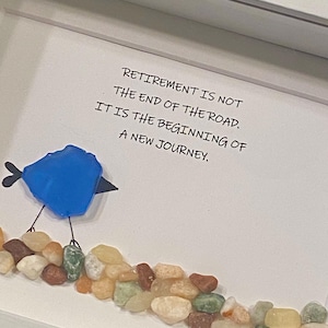 May include: Framed artwork with the text "RETIREMENT IS NOT THE END OF THE ROAD. IT IS THE BEGINNING OF A NEW JOURNEY." A blue bird made of glass sits above a row of colorful pebbles.