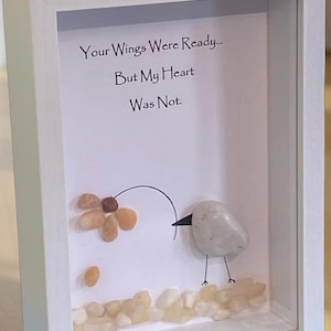 May include: A framed artwork with the text "Your Wings Were Ready... But My Heart Was Not." A pebble art bird is reaching for a flower made of pebbles. The artwork is in a white frame.