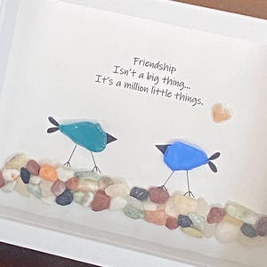 May include: A framed wall art piece featuring two birds made of teal and blue stones perched on a line of multicolored pebbles. The text reads: Friendship Isn't a big thing... It's a million little things. A small heart-shaped stone is nearby.