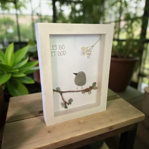 Könnte beinhalten: Weiß gerahmtes Kunstwerk mit einem Kieselkunst-Vogel, der auf einem Ast sitzt. Das Kunstwerk enthält den Text "LET GO ET GOD" und kleine grüne Kieselblätter. Der Hintergrund ist ein weiches Weiß.
