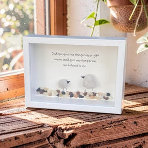 May include: A white framed art piece with a glass front. Inside, two pebble birds perch on a bed of small stones. The text reads, "Dad, you gave me the greatest gift anyone could give another person, you believed in me."