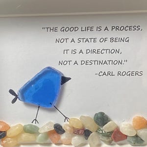 Peut inclure: Œuvre d'art encadrée représentant un oiseau bleu en verre avec des pattes et un bec noirs, marchant au-dessus d'une rangée de galets colorés. L'œuvre comprend la citation "LA BONNE VIE EST UN PROCESSUS, PAS UN ÉTAT D'ÊTRE, C'EST UNE DIRECTION, PAS UNE DESTINATION." de Carl Rogers.
