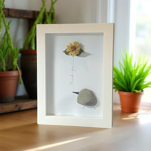 May include: White framed art piece featuring a dried yellow flower and a stone bird with the handwritten text "thank you". The artwork is displayed on a wooden surface, with plants in the background.