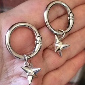 Può includere: Una coppia di anelli circolari in metallo color argento con chiusura a molla, ciascuno dotato di un piccolo ciondolo tridimensionale a forma di stella con quattro punte acuminate che si estendono da un punto centrale riflettente.