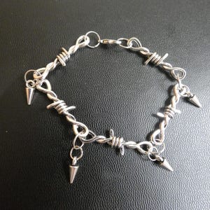 May include: A silver-colored bracelet with a barbed wire design. The bracelet features multiple loops of wire and small, cone-shaped spikes hanging from the loops. The bracelet is secured with a clasp.