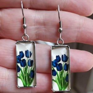 Könnte beinhalten: Ein Paar rechteckige Ohrhänger mit einem silberfarbenen Metallrahmen und einem Glaseinsatz. Das Design zeigt eine botanische Illustration von dunkelblauen Tulpen mit leuchtend grünen Stielen und Blättern auf einem cremefarbenen Hintergrund.