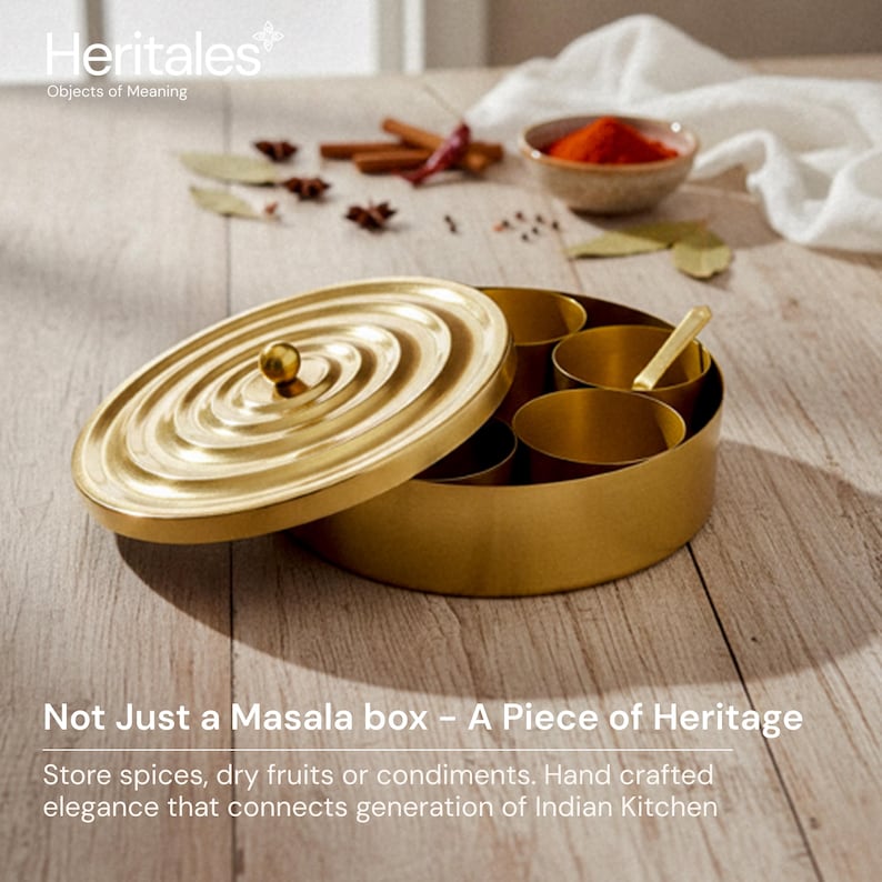 May include: A gold-colored masala box with a lid, featuring a spiral design. The box is open, revealing several compartments and a small spoon. Spices and condiments are visible in the background. Text on the image reads: "Not Just a Masala box - A Piece of Heritage."