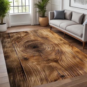 Puede incluir: Una alfombra rectangular con diseño de vetas de madera. La alfombra presenta un patrón detallado de círculos concéntricos y vetas lineales en tonos marrones y beige, imitando el aspecto de una sección transversal de un tronco de árbol. La alfombra está colocada sobre un suelo de madera.