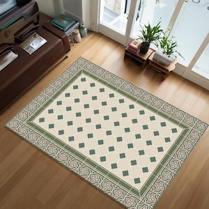 May include: A rectangular rug with a cream-coloured central design of dark green squares. The rug has a decorative border with floral and geometric patterns in shades of green, grey, and cream. The rug is placed on a wooden floor.