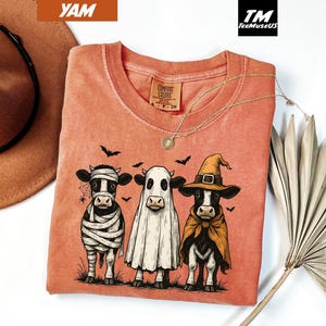 May include: A coral-colored Comfort Colors t-shirt featuring a Halloween-themed graphic of three cows dressed as a mummy, ghost, and witch. The design includes bats and a spiderweb. A gold necklace is draped on the shirt.