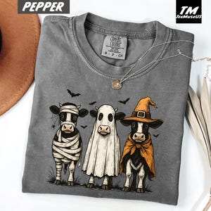 May include: A gray Comfort Colors t-shirt featuring three cows dressed in Halloween costumes: a mummy, a ghost, and a witch. The graphic includes bats and a spiderweb. The shirt has a crew neck and short sleeves.