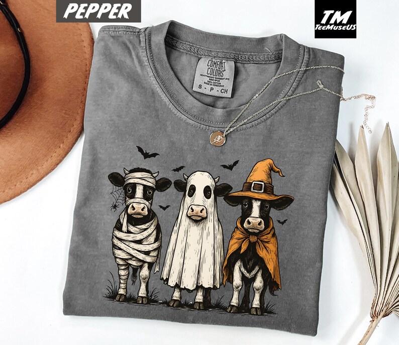 May include: A gray Comfort Colors t-shirt featuring three cows dressed in Halloween costumes: a mummy, a ghost, and a witch. The graphic includes bats and a spiderweb. The shirt has a crew neck and short sleeves.