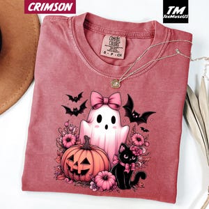 Pode incluir: Camiseta carmesim com um gr&aacute;fico tem&aacute;tico de Halloween. O design inclui um fantasma com um la&ccedil;o rosa, uma ab&oacute;bora esculpida, um gato preto e morcegos. A camisa tem gola redonda e uma etiqueta Comfort Colors.