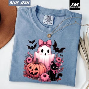 Pode incluir: Camiseta Comfort Colors azul jeans com um design de Halloween. O design inclui um fantasma rosa com um la&ccedil;o, uma ab&oacute;bora, um gato preto e morcegos. A camiseta tamb&eacute;m tem um colar dourado.