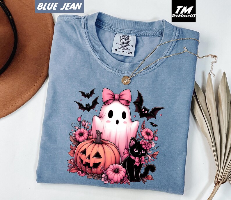 Pode incluir: Camiseta Comfort Colors azul jeans com um design de Halloween. O design inclui um fantasma rosa com um la&ccedil;o, uma ab&oacute;bora, um gato preto e morcegos. A camiseta tamb&eacute;m tem um colar dourado.