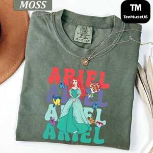 May include: Moss green t-shirt featuring the word "ARIEL" in red, purple, and teal, with a graphic of Ariel from The Little Mermaid. The shirt has a crew neck and short sleeves. A gold necklace is draped across the shirt.