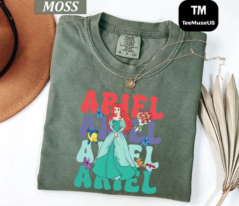 May include: Moss green t-shirt featuring the word "ARIEL" in red, purple, and teal, with a graphic of Ariel from The Little Mermaid. The shirt has a crew neck and short sleeves. A gold necklace is draped across the shirt.